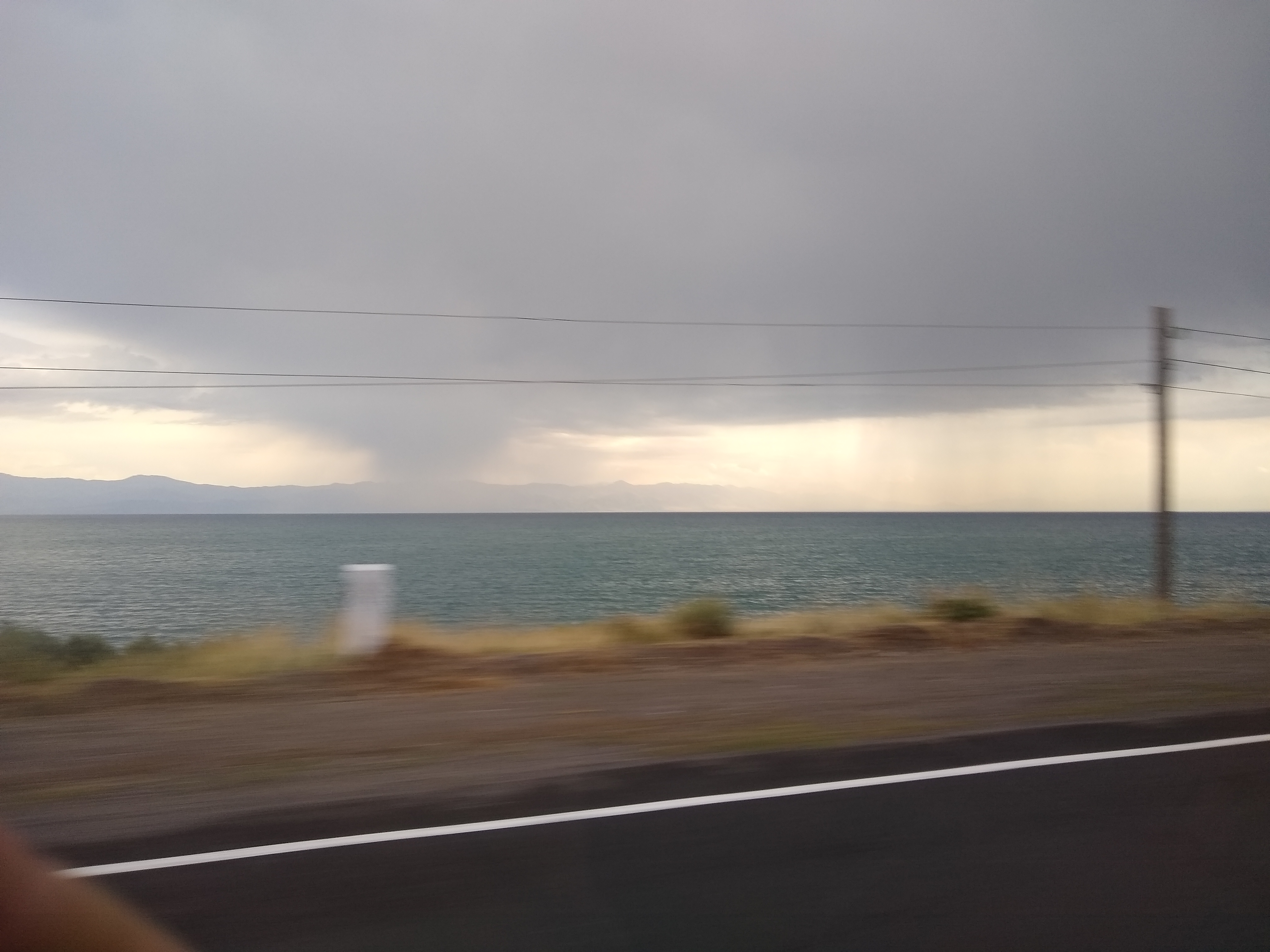 Blurry photo taken from a moving vehicle of a large lake with a dark grey storm cloud overhead