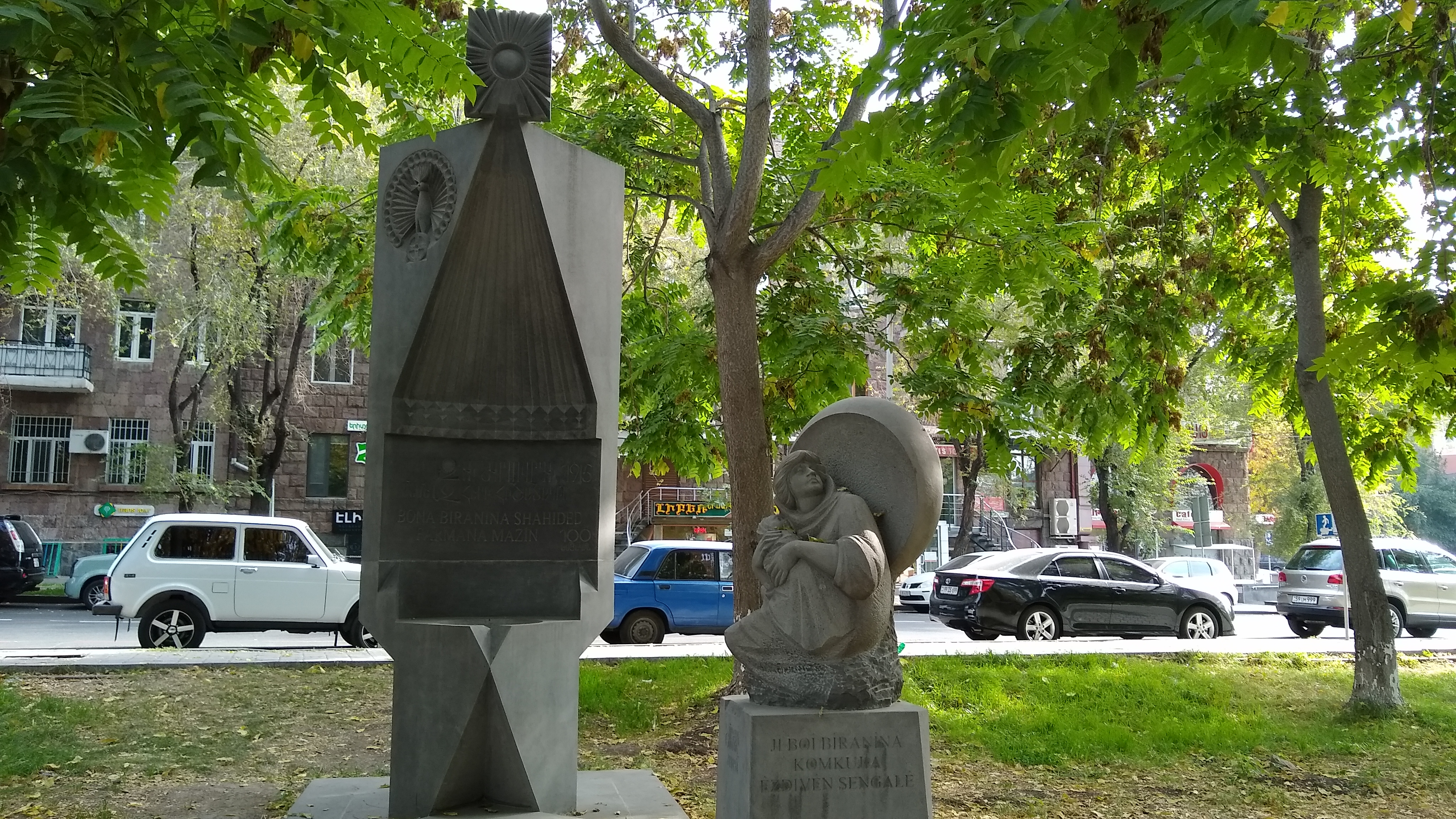 A photo of a masonary monument, including a statue of someone holding a child, with trees and a street in the background.
