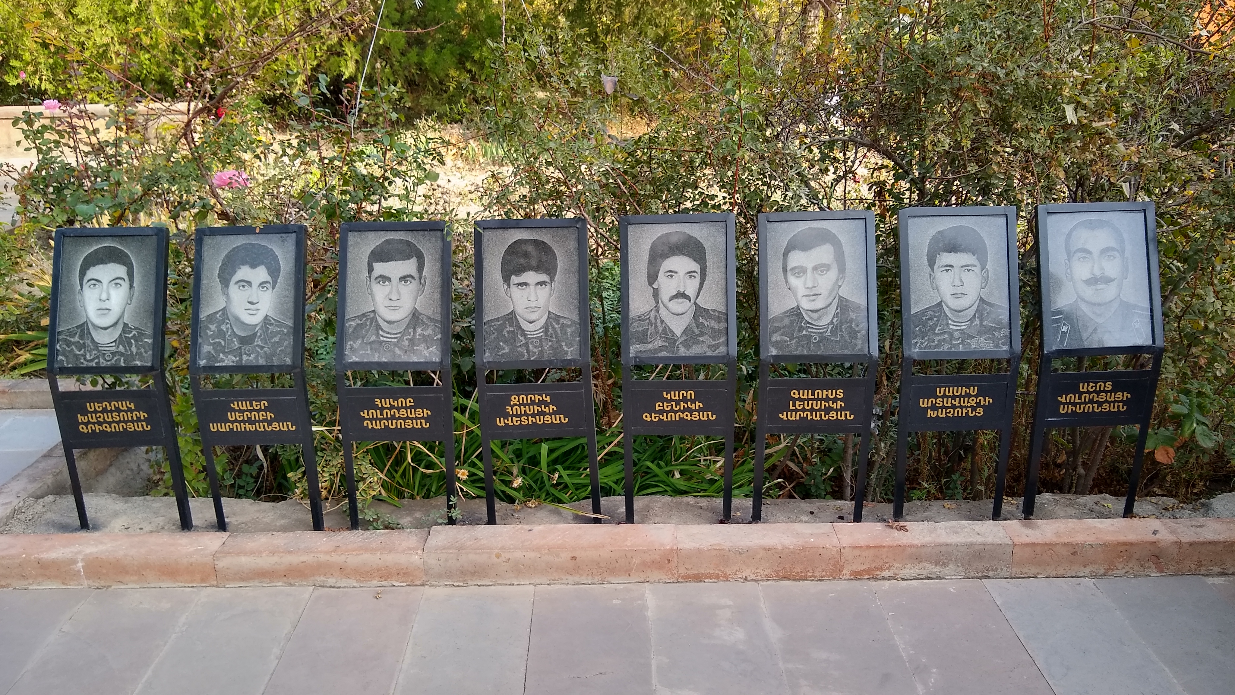 Photos of eight men in military fatigues on plaques, with Armenian writing below them, with a hedge in the background.