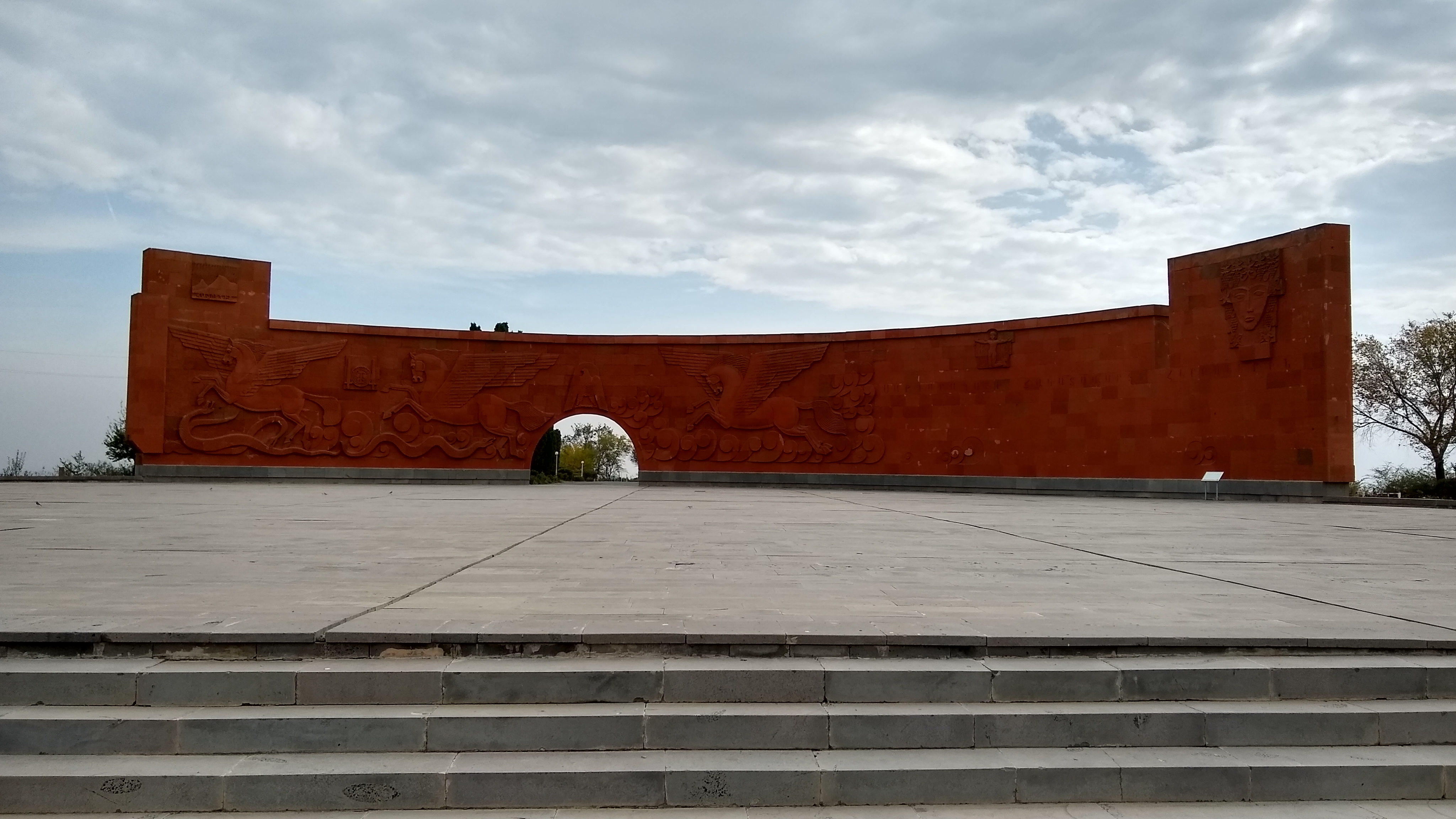 A photo of a large stone courtyard with a relief in the background carved into red stone featuring winged horses.